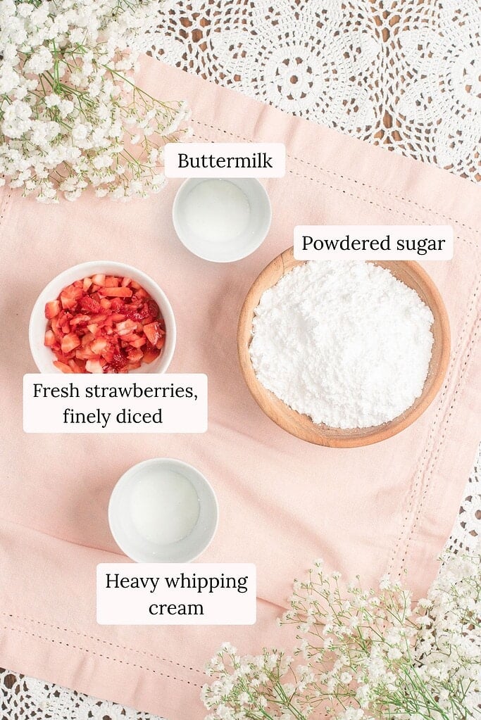 Overhead flat lay of glaze ingredients including a small bowl of buttermilk, a bowl of finely diced fresh strawberries, a wooden bowl of powdered sugar, and a bowl of heavy whipping cream arranged on pink linen with baby's breath