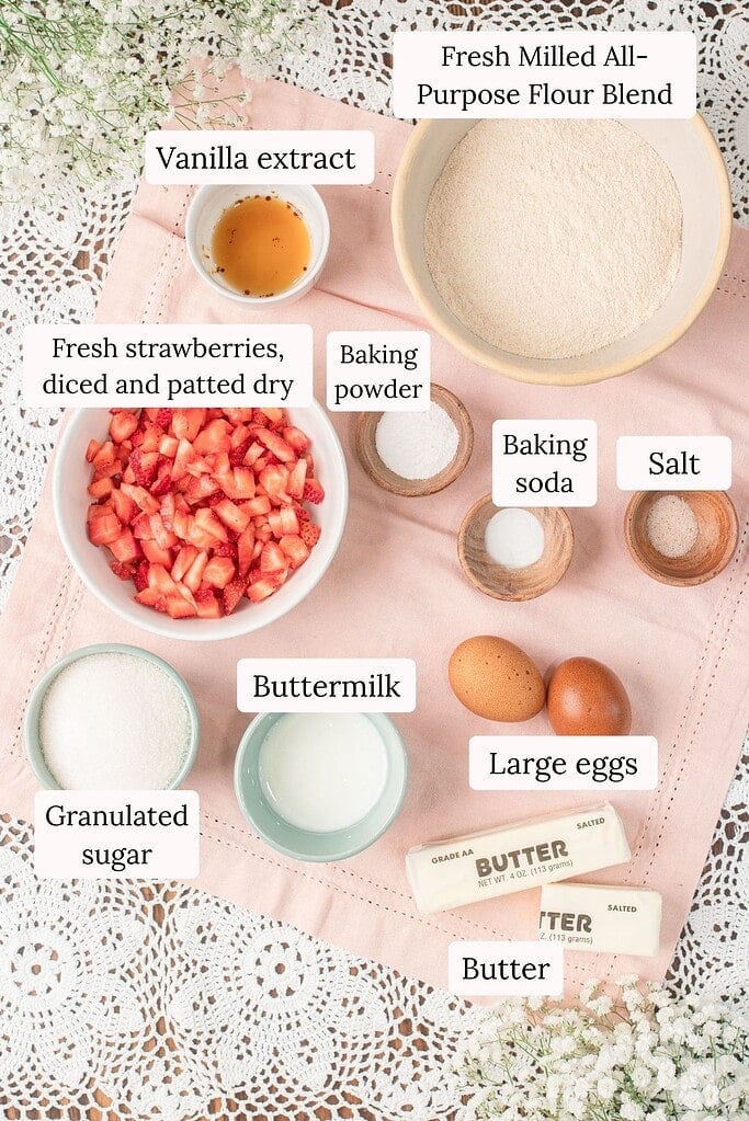 Overhead flat lay of ingredients for Fresh Milled Flour Strawberry Bread including a bowl of fresh milled all-purpose flour blend, diced strawberries patted dry, buttermilk, eggs, butter, granulated sugar, vanilla extract, and small bowls of baking powder, baking soda, and salt