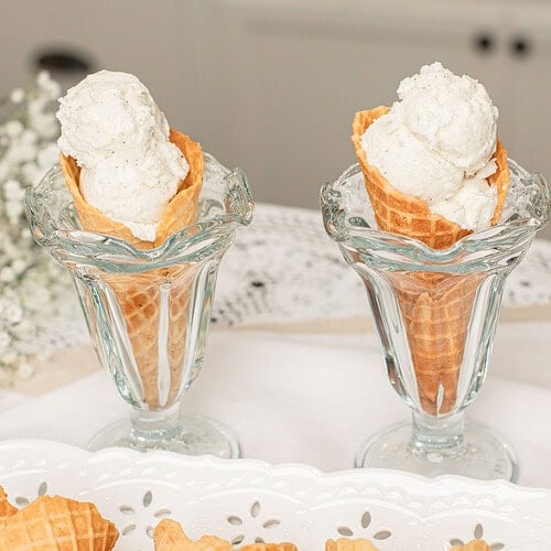 Homemade fresh milled flour waffle cones filled with scoops of vanilla ice cream in vintage glass dessert cups, with additional crisp waffle cones arranged on a white serving tray.