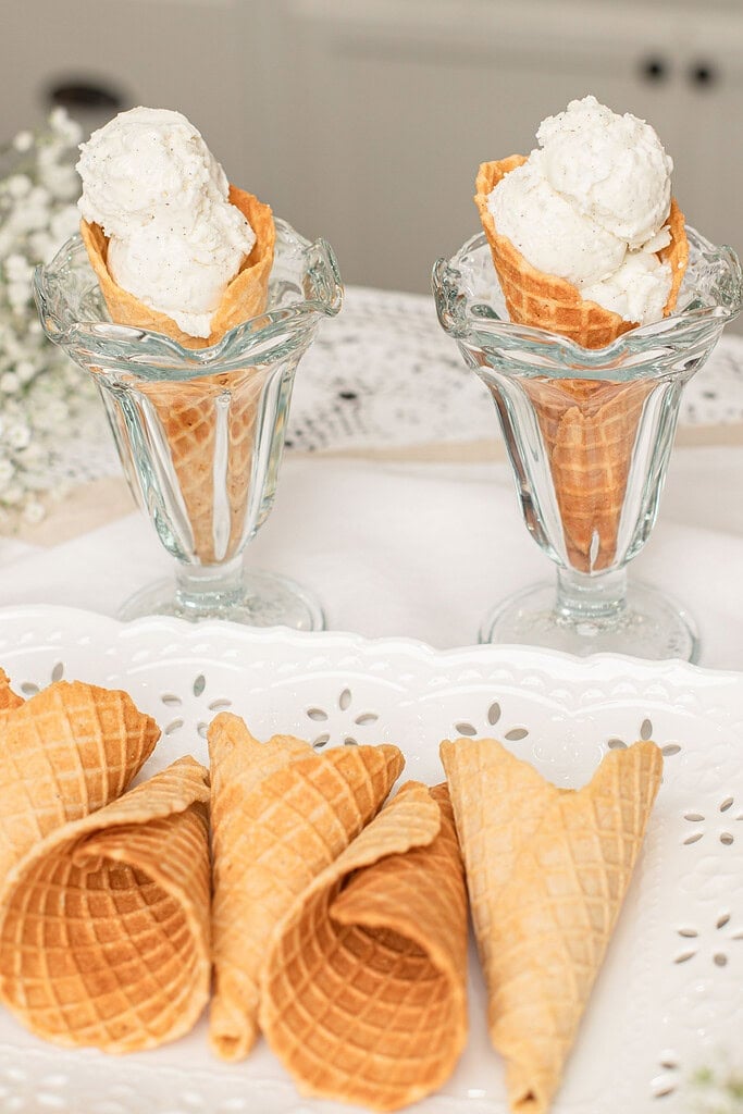 Homemade fresh milled flour waffle cones filled with scoops of vanilla ice cream in vintage glass dessert cups, with additional crisp waffle cones arranged on a white serving tray.