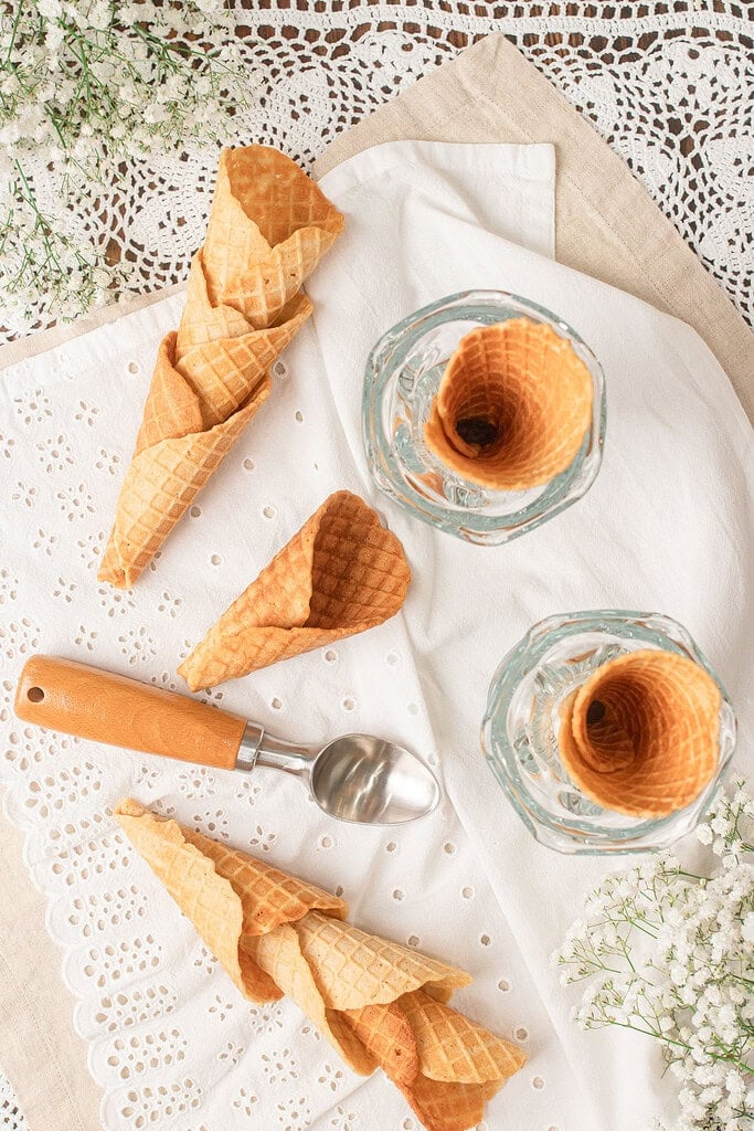 Overhead view of homemade fresh milled flour waffle cones arranged on a white linen cloth with vintage glass dessert cups and an ice cream scoop, ready for serving.
