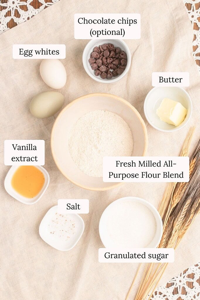 Flat lay of ingredients for fresh milled flour waffle cones including fresh milled all-purpose flour blend, granulated sugar, butter, egg whites, vanilla extract, salt, and optional chocolate chips arranged on a cream linen cloth with wheat stalks.