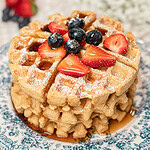 Stack of golden waffles made with freshly milled flour on a blue and white floral plate, topped with powdered sugar, fresh strawberries, and blueberries, with more berries blurred in the background.
