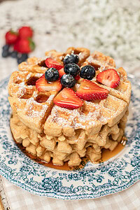 Stack of golden waffles made with freshly milled flour on a blue and white floral plate, topped with powdered sugar, fresh strawberries, and blueberries, with more berries blurred in the background.