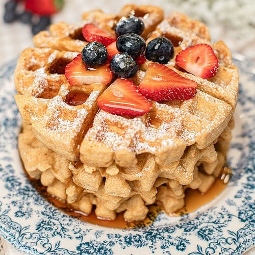 Stack of golden waffles made with freshly milled flour on a blue and white floral plate, topped with powdered sugar, fresh strawberries, and blueberries, with more berries blurred in the background.