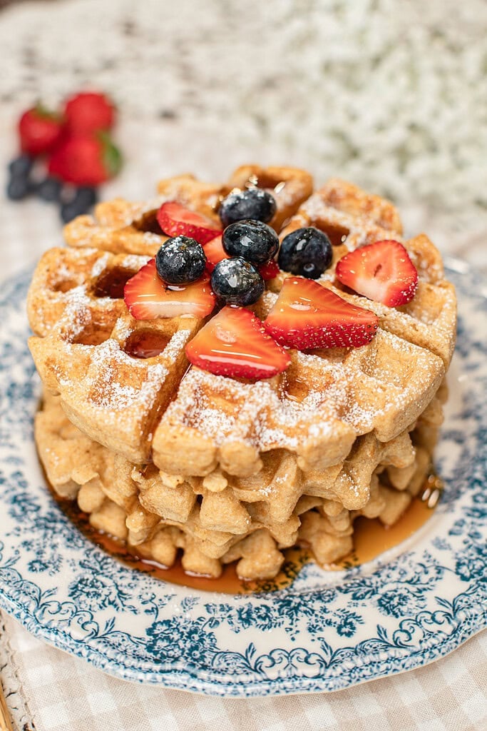 Stack of golden waffles made with freshly milled flour on a blue and white floral plate, topped with powdered sugar, fresh strawberries, and blueberries, with more berries blurred in the background.