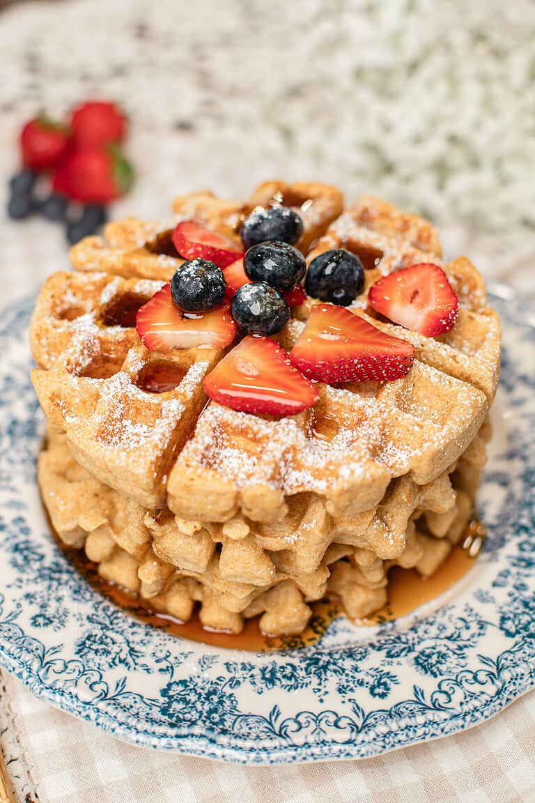 Stack of golden waffles made with freshly milled flour on a blue and white floral plate, topped with powdered sugar, fresh strawberries, and blueberries, with more berries blurred in the background.