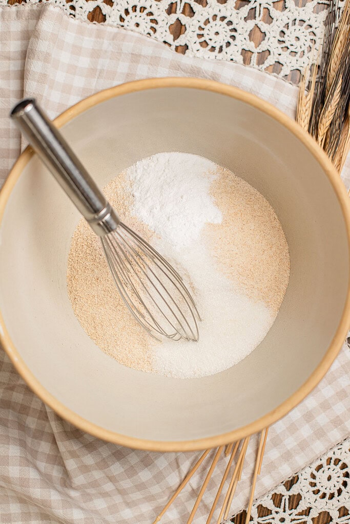 Overhead view of freshly milled flour and sugar in a beige mixing bowl with a whisk resting inside, dried wheat stalks visible beside the bowl on a checked cloth.