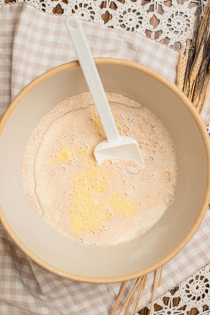 Overhead view of eggs and melted butter poured over freshly milled flour in a mixing bowl, not yet stirred together, with a spatula resting inside and dried wheat stalks nearby.