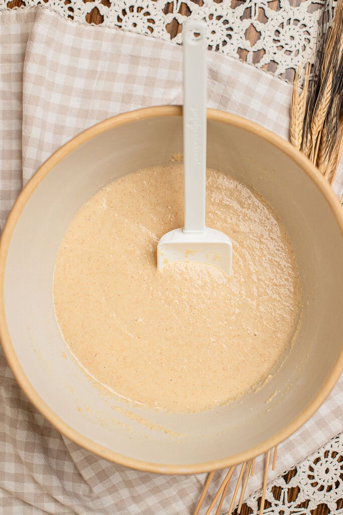 Overhead view of smooth freshly milled flour waffle batter mixed in a beige bowl with a spatula resting inside, surrounded by dried wheat stalks and a checked cloth.