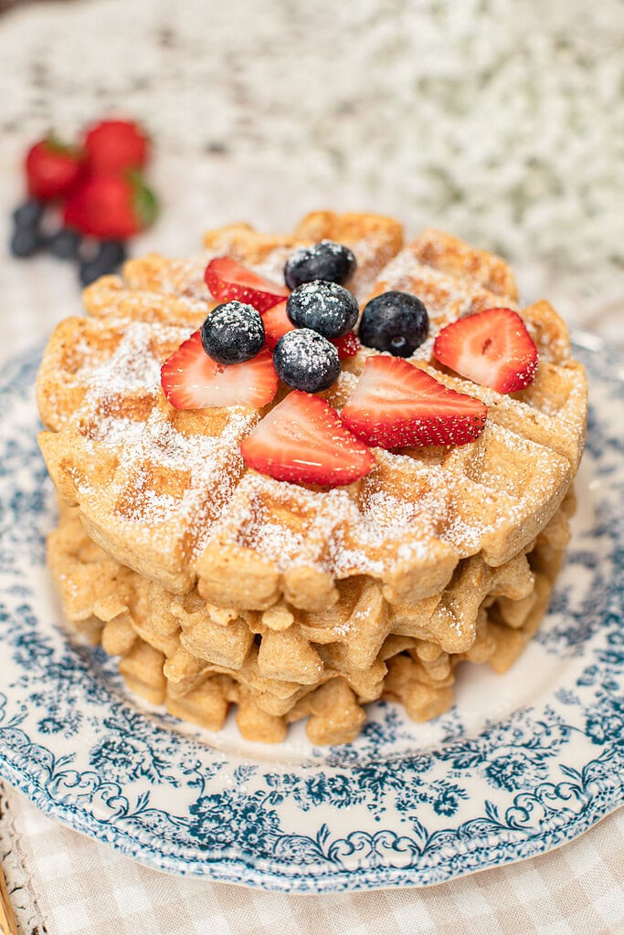 Close-up of a stack of freshly milled flour waffles dusted with powdered sugar and topped with fresh strawberries and blueberries, with more berries blurred in the background.