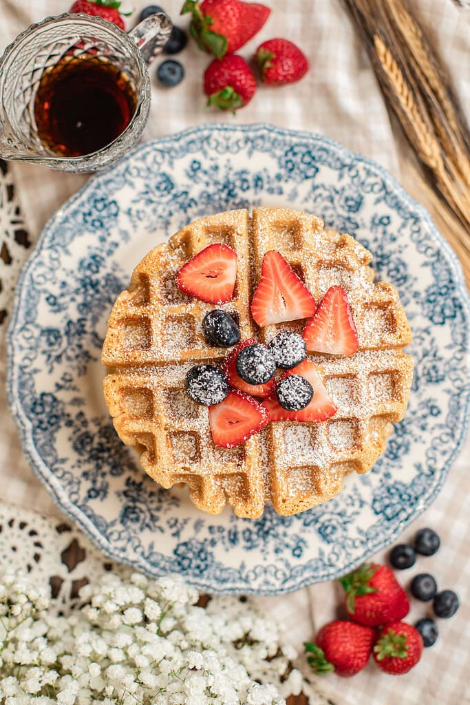 Overhead view of a single golden waffle made with freshly milled flour on a blue and white floral plate, topped with powdered sugar, strawberries, and blueberries, with a small glass of maple syrup and dried wheat stalks nearby.
