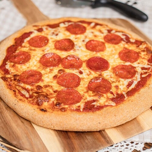 A whole baked pepperoni pizza on freshly milled flour dough, set on a wooden board next to a pizza cutter