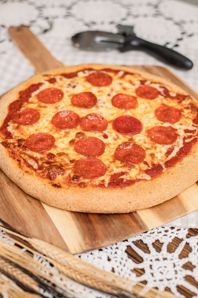 A whole baked pepperoni pizza on freshly milled flour dough, set on a wooden board next to a pizza cutter