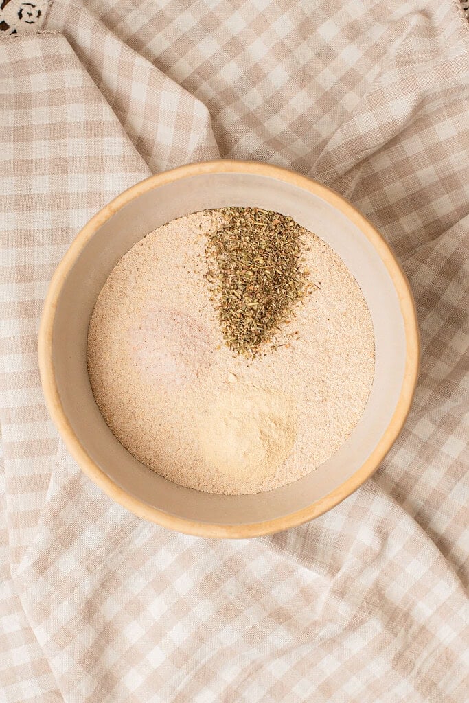 Dried oregano, garlic powder, and seasonings sprinkled over freshly milled flour in a bow