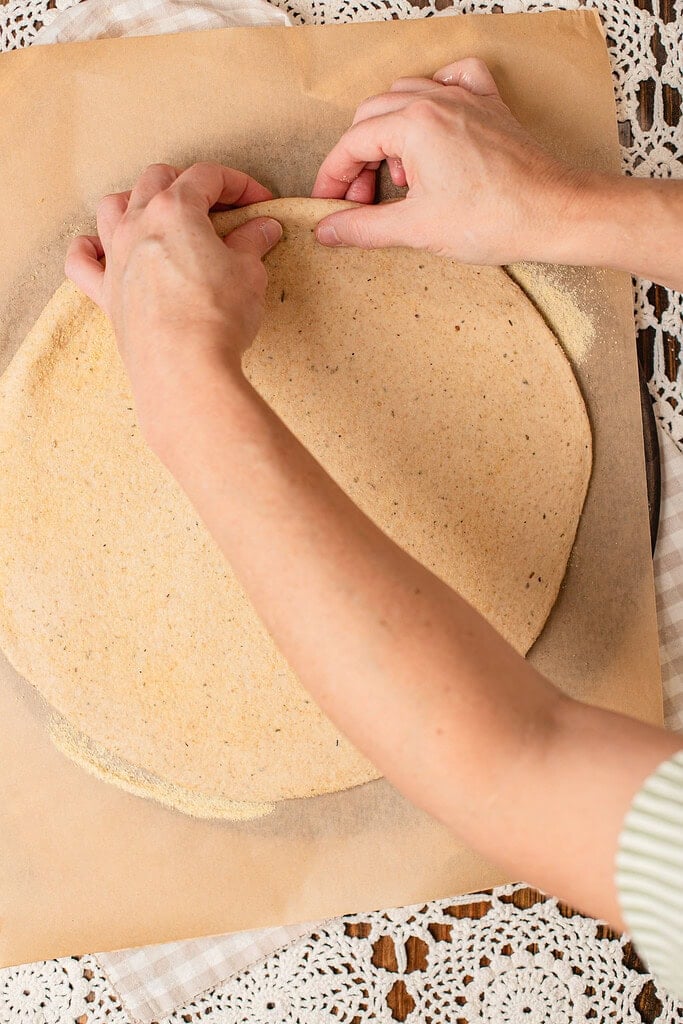 Hands stretching freshly milled flour pizza dough into a round crust on cornmeal-dusted parchment paper