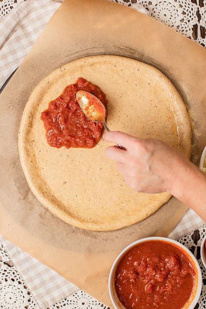 A spoon spreading marinara sauce over a stretched round of freshly milled flour pizza dough