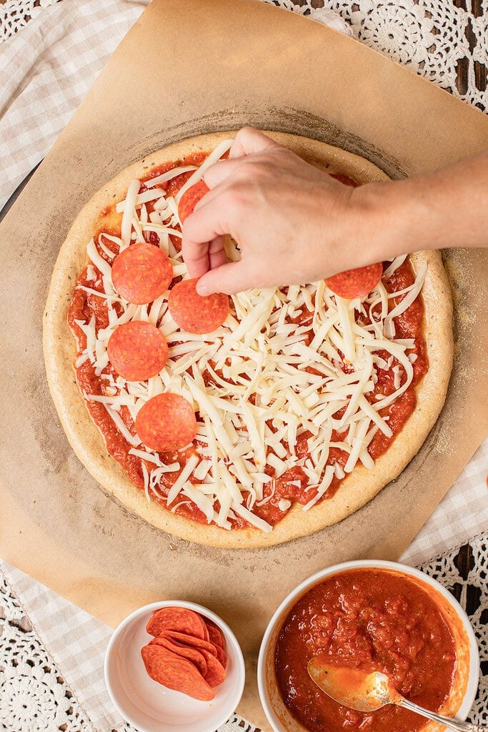 A hand placing pepperoni over shredded mozzarella on a sauced freshly milled flour pizza crust