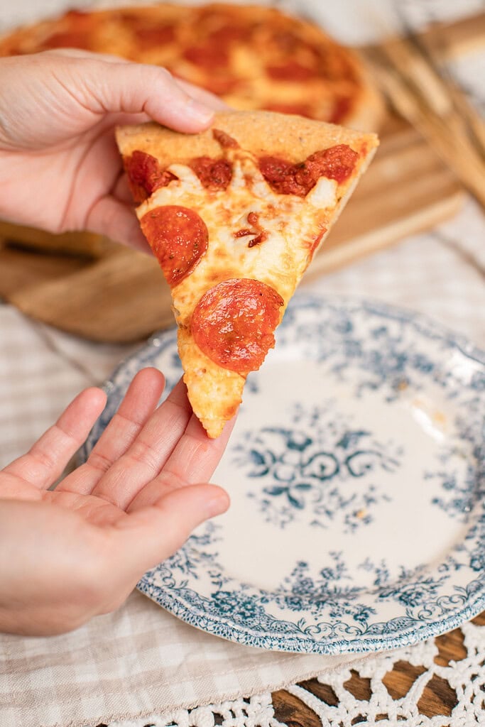 A slice of pepperoni pizza on freshly milled flour dough held above a blue floral plate, showing the crust and toppings up close