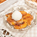 A slice of peach cobbler with sourdough topped with vanilla ice cream on a vintage plate, with the full baking dish and a second serving visible in the background.