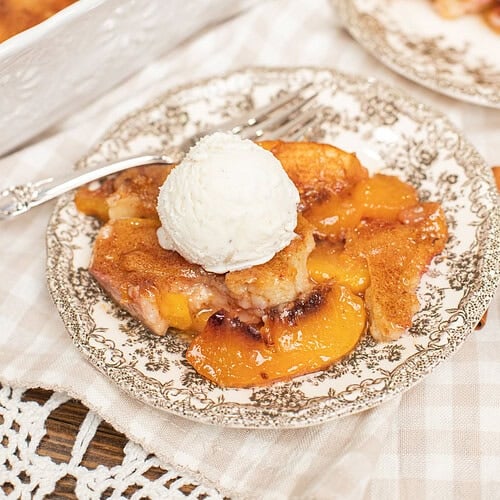 A slice of peach cobbler with sourdough topped with vanilla ice cream on a vintage plate, with the full baking dish and a second serving visible in the background.