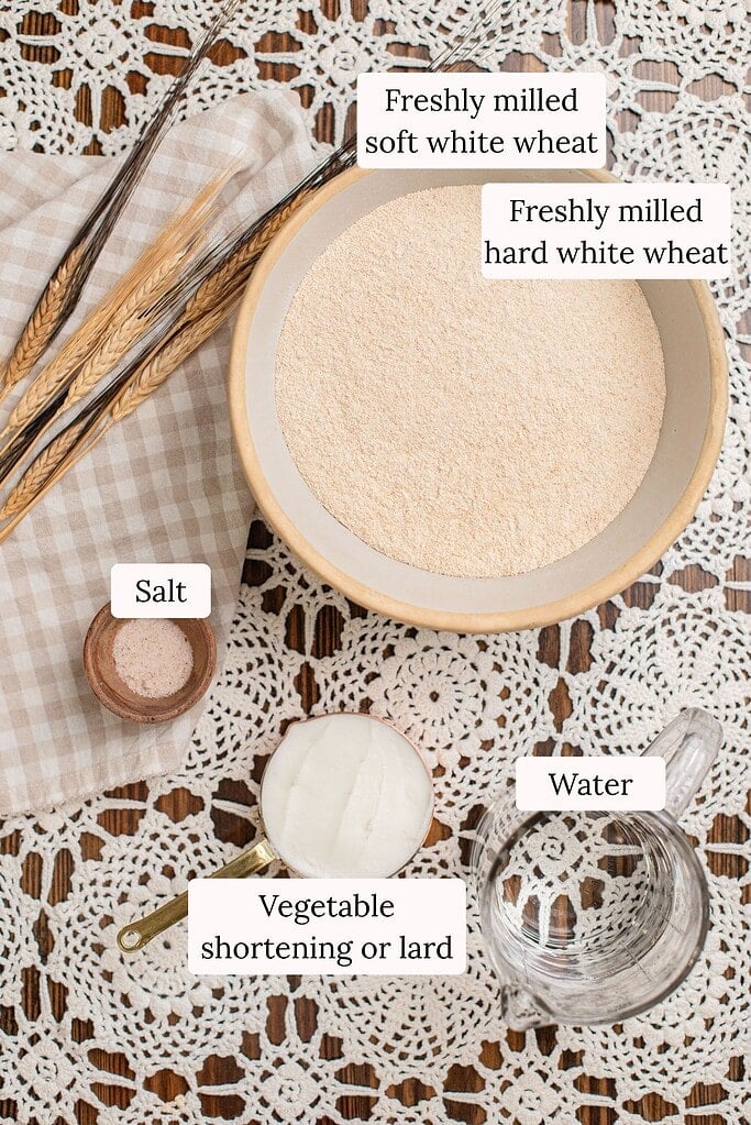 Freshly milled flour in a bowl with salt, oil, and water measured out beside it, wheat stalks resting on a checked cloth