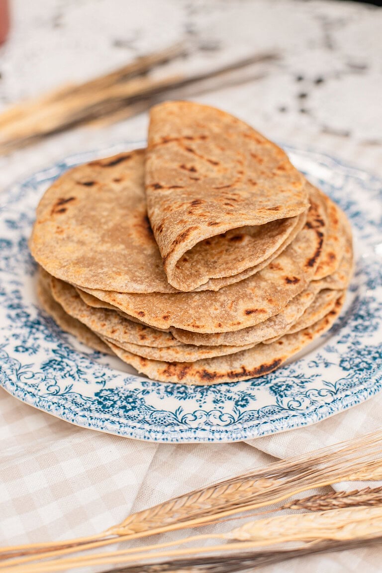 A stack of folded tortillas made with freshly milled flour piled on a blue floral plate, char marks visible on top