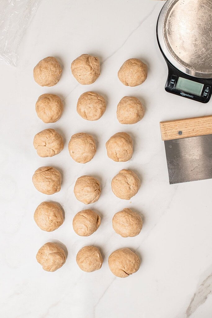 Rows of small dough balls portioned out on a marble counter next to a kitchen scale and dough cutter, ready to roll into tortillas