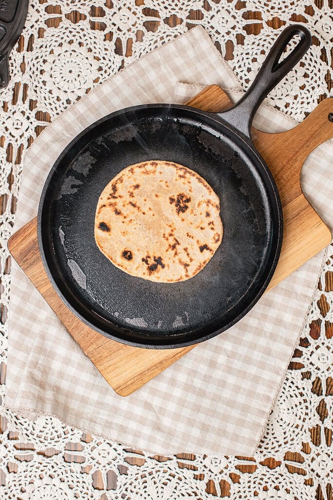 A freshly milled flour tortilla browning in a cast iron skillet with charred spots forming on the surface