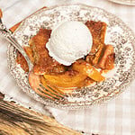 A slice of peach cobbler made with freshly milled flour topped with vanilla ice cream on a vintage plate, cast iron skillet and a second serving blurred in the background.