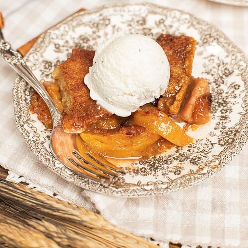 A slice of peach cobbler made with freshly milled flour topped with vanilla ice cream on a vintage plate, cast iron skillet and a second serving blurred in the background.
