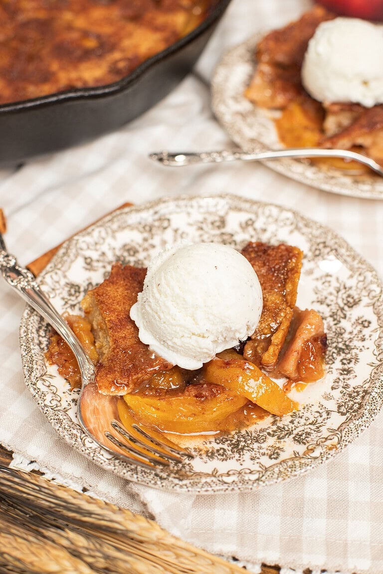 A slice of peach cobbler made with freshly milled flour topped with vanilla ice cream on a vintage plate, cast iron skillet and a second serving blurred in the background.