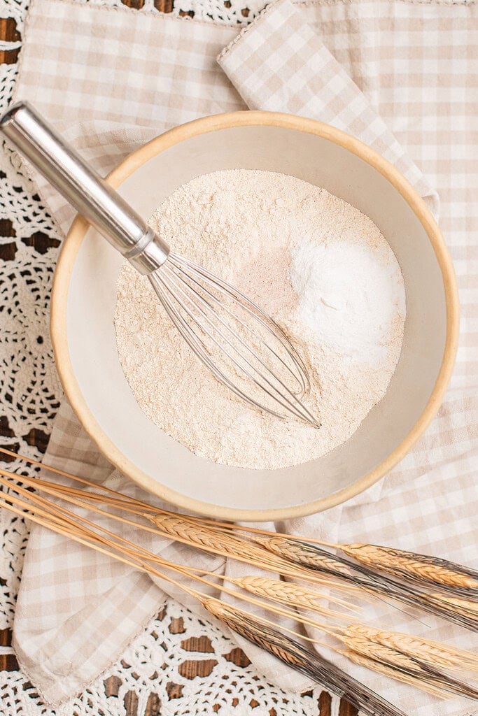 Freshly milled flour in a bowl with a whisk, baking soda and salt piled to one side, wheat stalks laid across a checked cloth