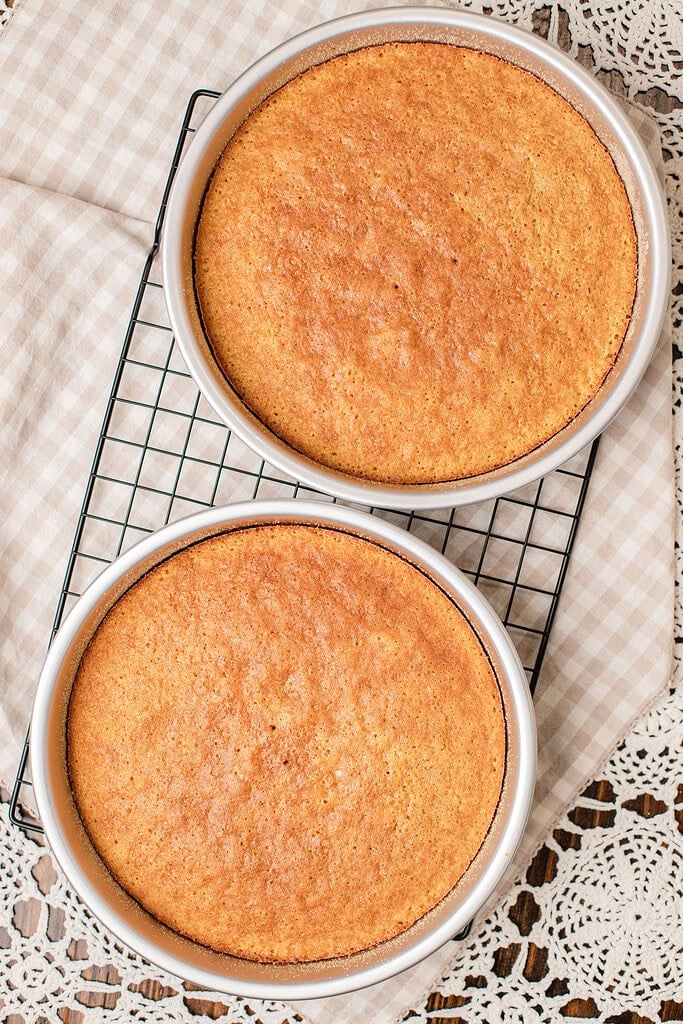 Two golden baked cake layers cooling in their pans on a wire rack, sugared crust visible on top