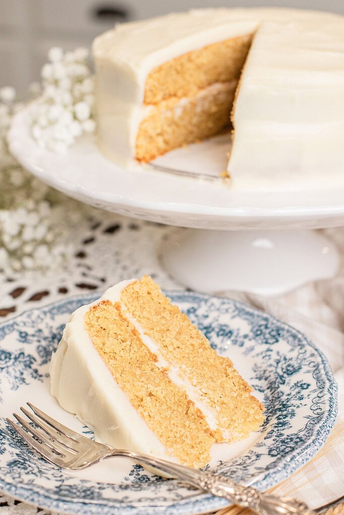 A slice of yellow cake with cream cheese frosting on a blue floral plate, fork resting beside it, whole cake visible in the background