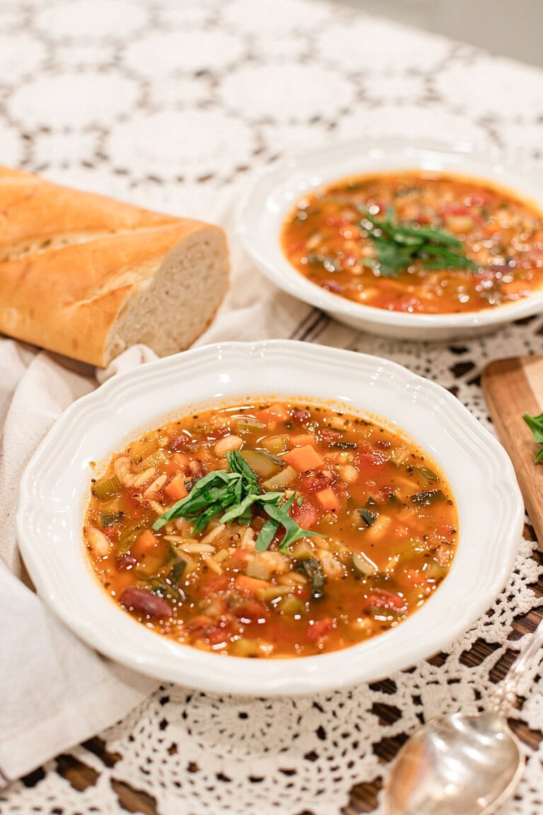 Two white scalloped bowls of homemade minestrone soup topped with fresh basil, served with a sliced baguette on a lace tablecloth